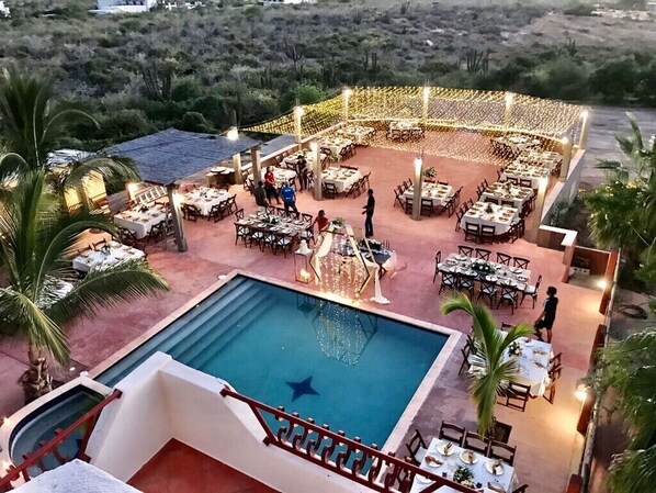 A poolside dining area with tables and chairs, surrounded by a string of lights.