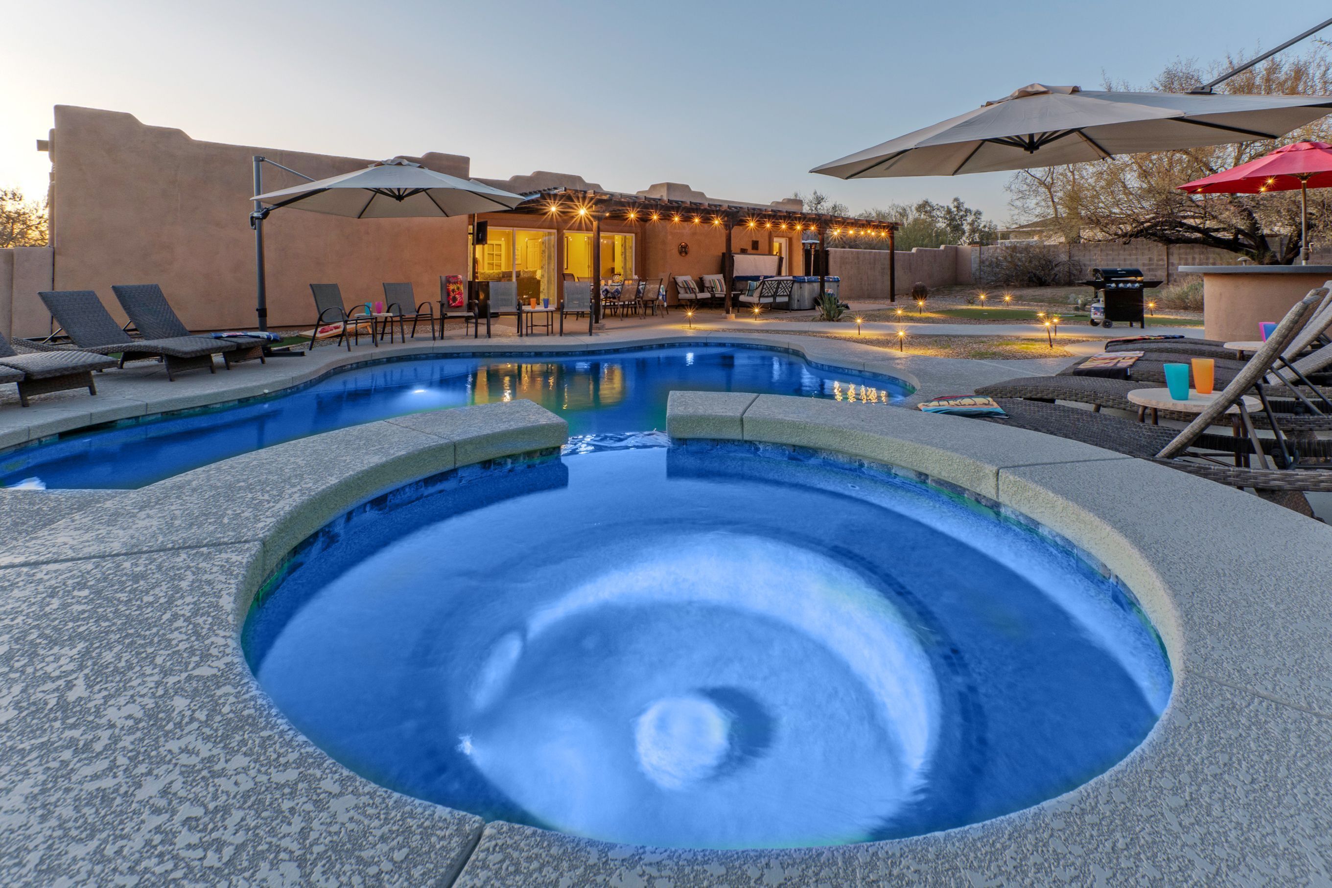 Una piscina con sillas de descanso y sombrillas, rodeada por un edificio con luces colgantes.