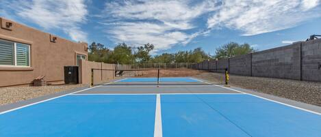An outdoor tennis court with a blue surface, surrounded by a low stone wall and a building with beige walls and a door.