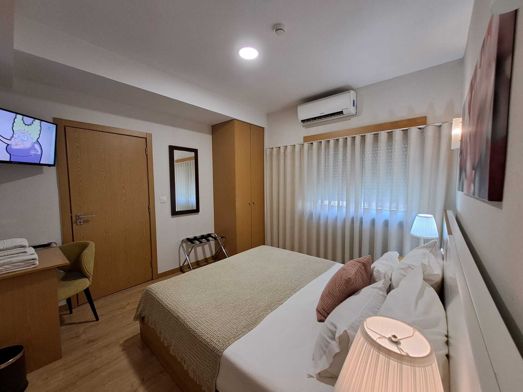 A hotel room with a bed, a desk, a chair, a television, an air conditioning unit, and a window with curtains.