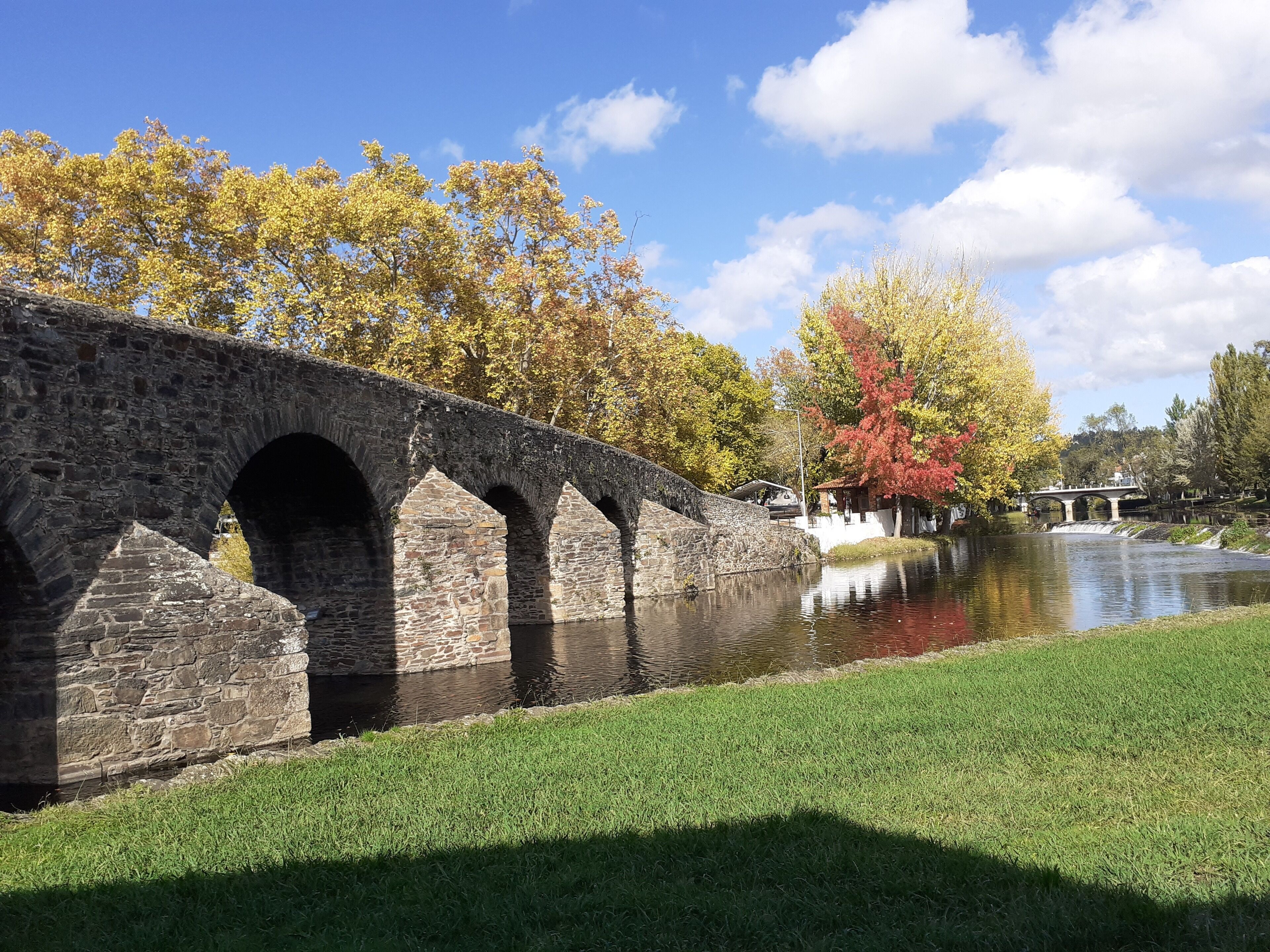 A stone bridge over a calm river, surrounded by grassy banks and autumn-colored trees.