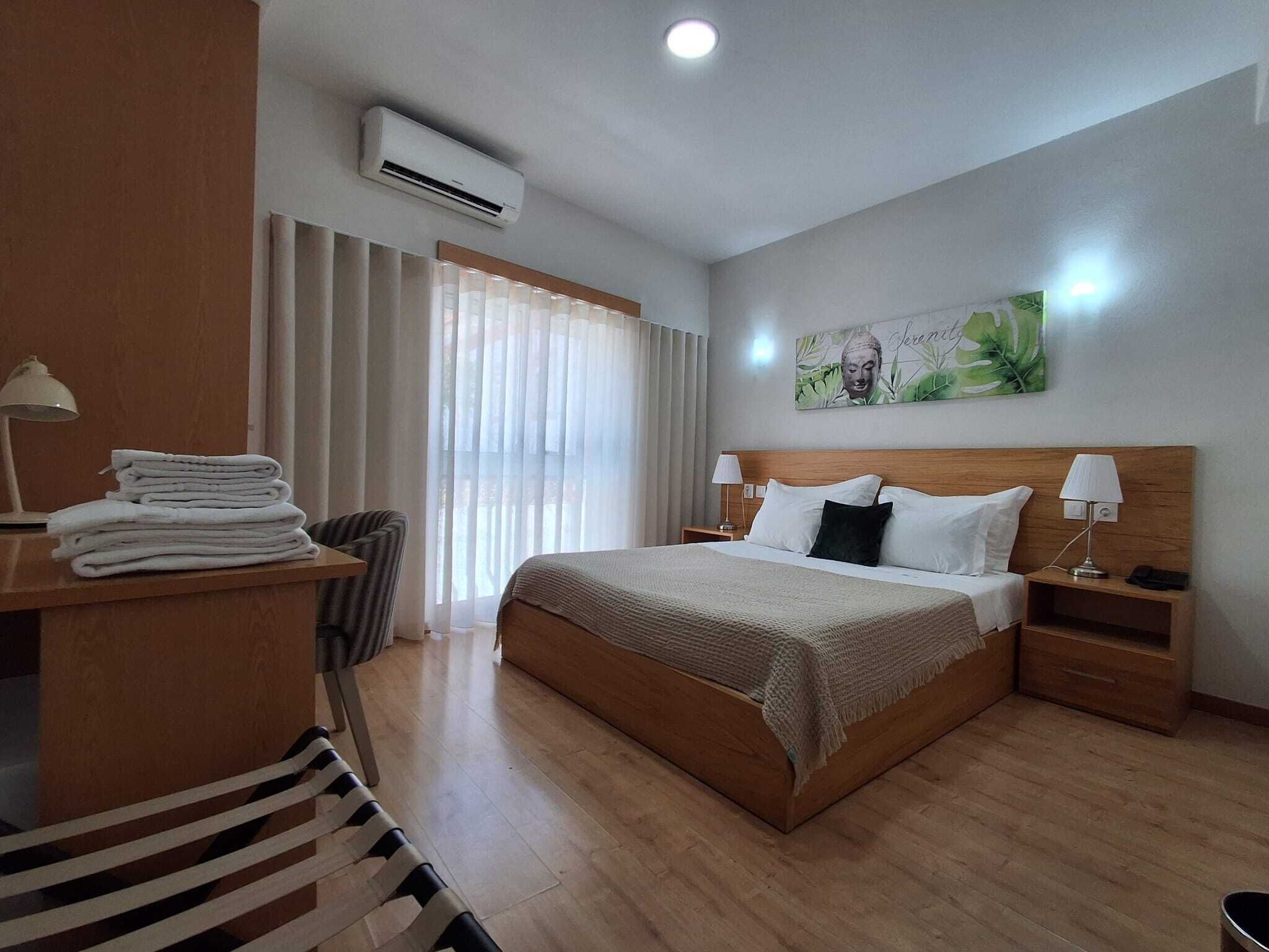 A hotel room with a bed, bedside tables, a desk with a lamp, a chair, and a window with curtains.
