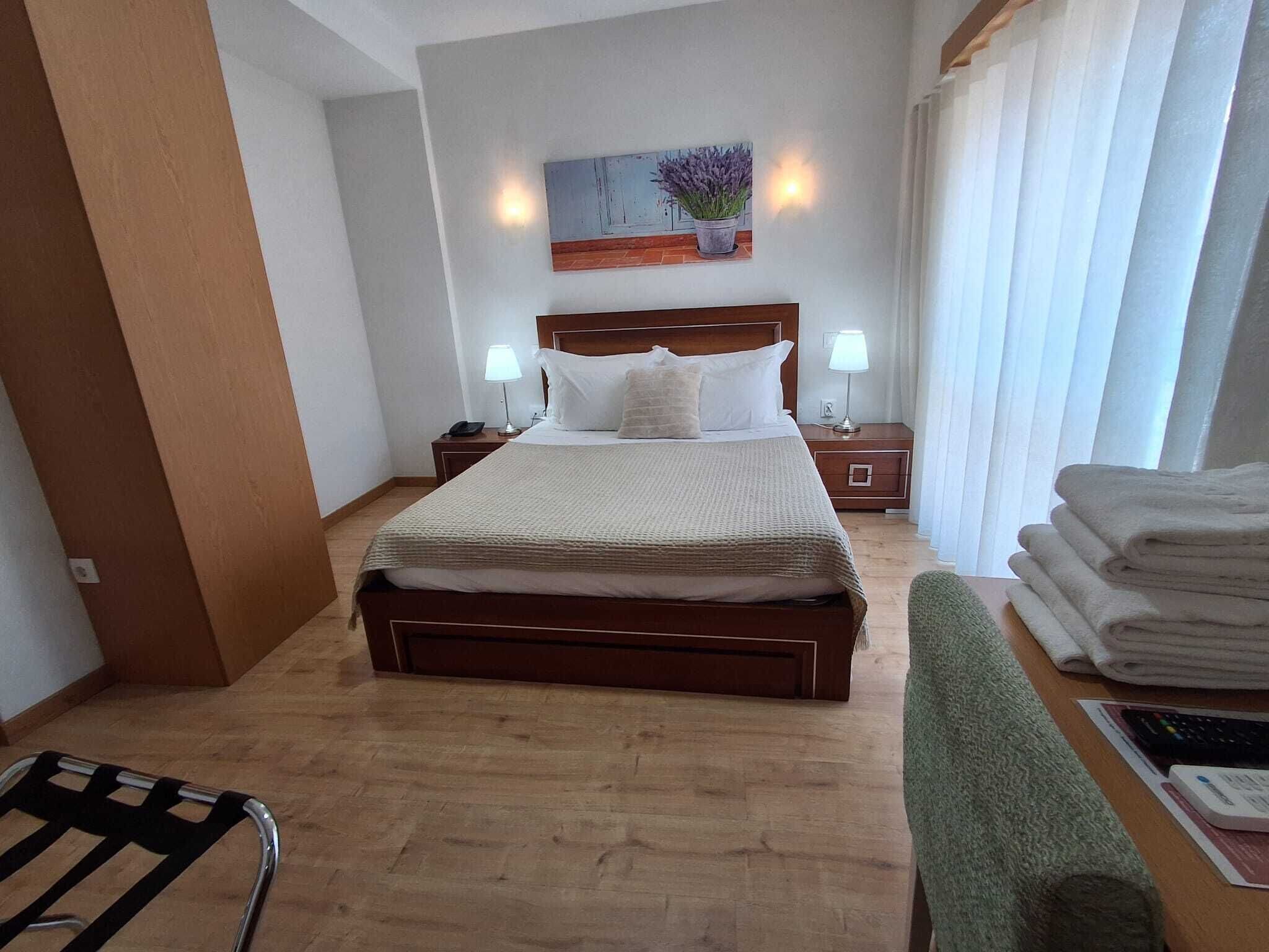 Habitación de hotel con cama de madera, dos mesitas de noche con lámparas, una obra de arte en la pared y un ventanal con cortinas.