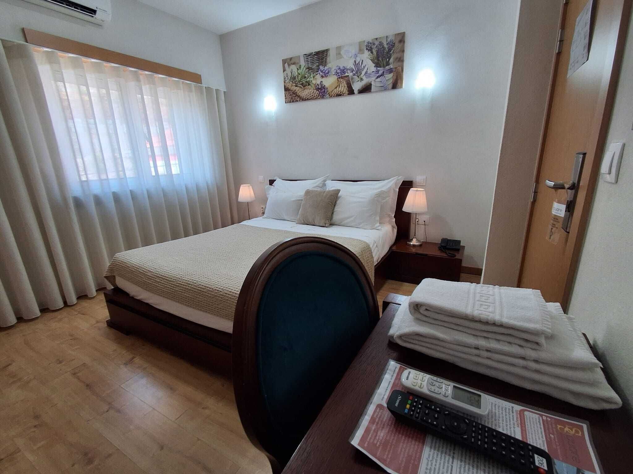 A hotel room with a bed, bedside lamps, a wooden nightstand, a window with curtains, and a wall-mounted picture.