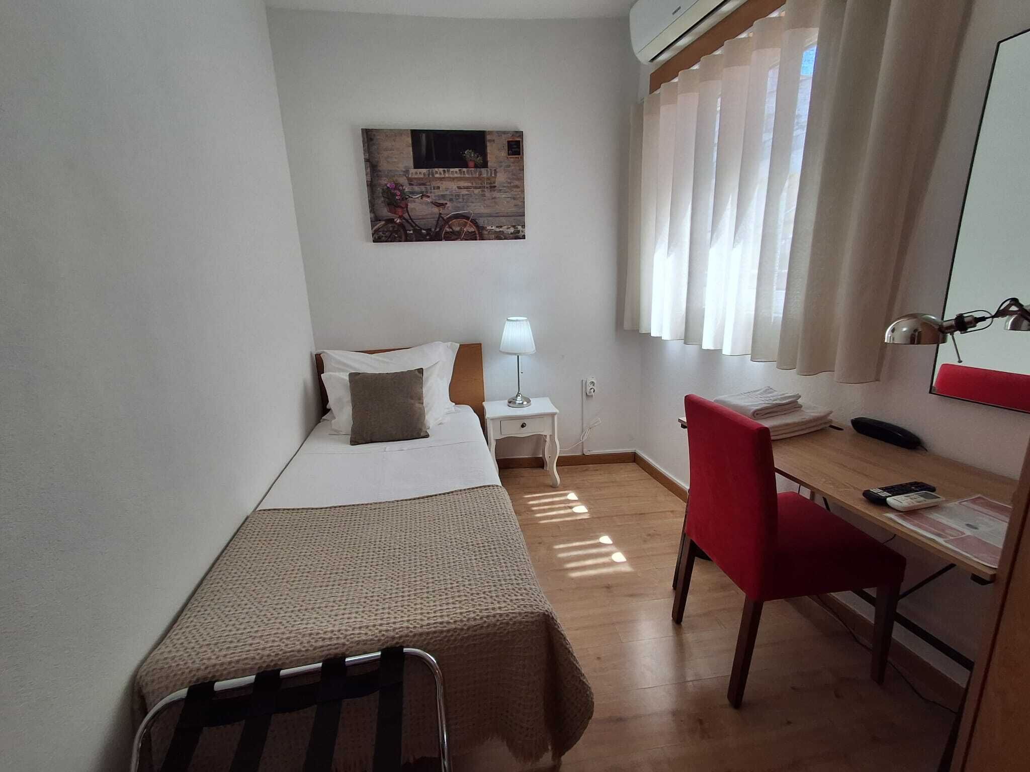 Une petite chambre d’hôtel, propre et bien rangée, avec un lit simple, un bureau avec une chaise, une lampe et une fenêtre avec des rideaux.