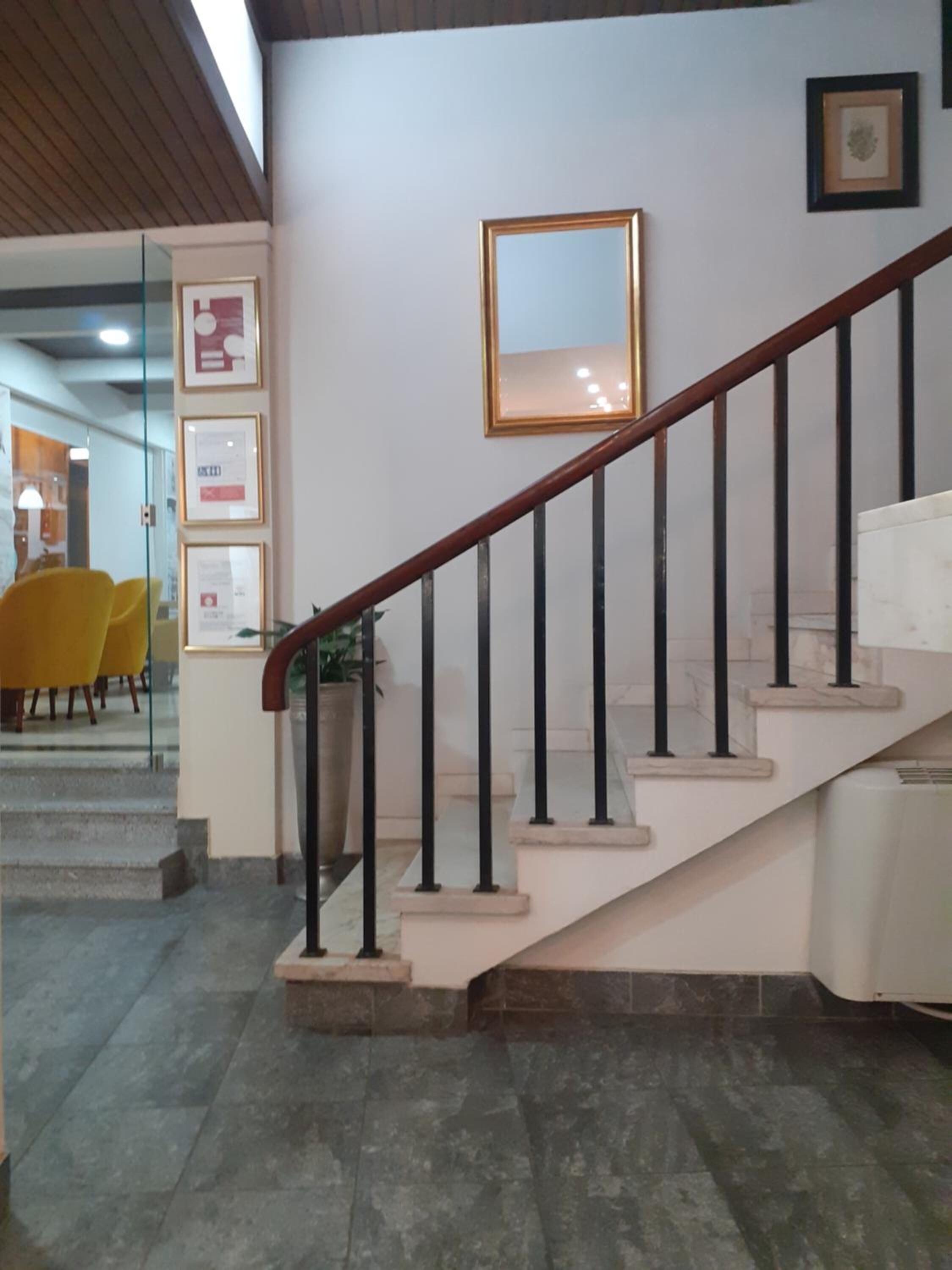 Una escalera con barandilla de madera y balaustradas negras, que conduce a un rellano con un espejo enmarco y diplomas en la pared.