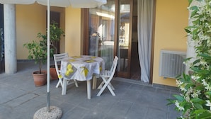 En udendørs terrasse med et bord og stole, en hvid parasol, plantekasser og en skydedør.