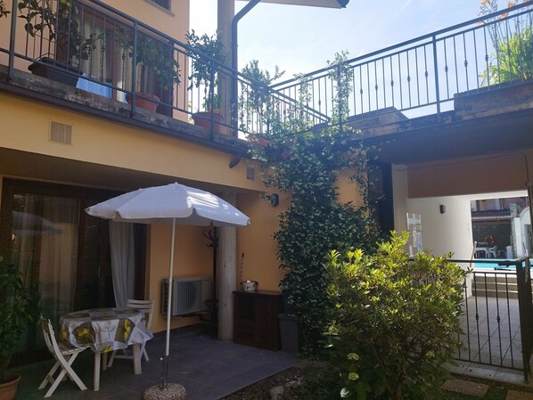 Un patio con una mesa y sillas bajo un paraguas blanco, rodeado de plantas en macetas y un pequeño jardín.