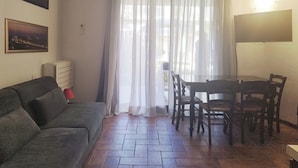 En stue med sofa, spisebord med stole, et vindue med lette gardiner og en vægmonteret aircondition.