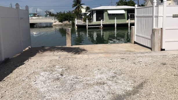 Un port de plaisance fermé, doté d’un quai en béton qui s’avance dans une eau calme, et entouré d’une clôture blanche et de palmiers.