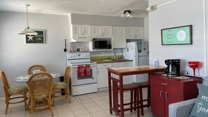 Une cuisine compacte avec des meubles blancs, un four à micro-ondes, une cuisinière et un réfrigérateur. On y trouve une table à manger avec des chaises, ainsi qu’un petit meuble rouge contenant une machine à café.