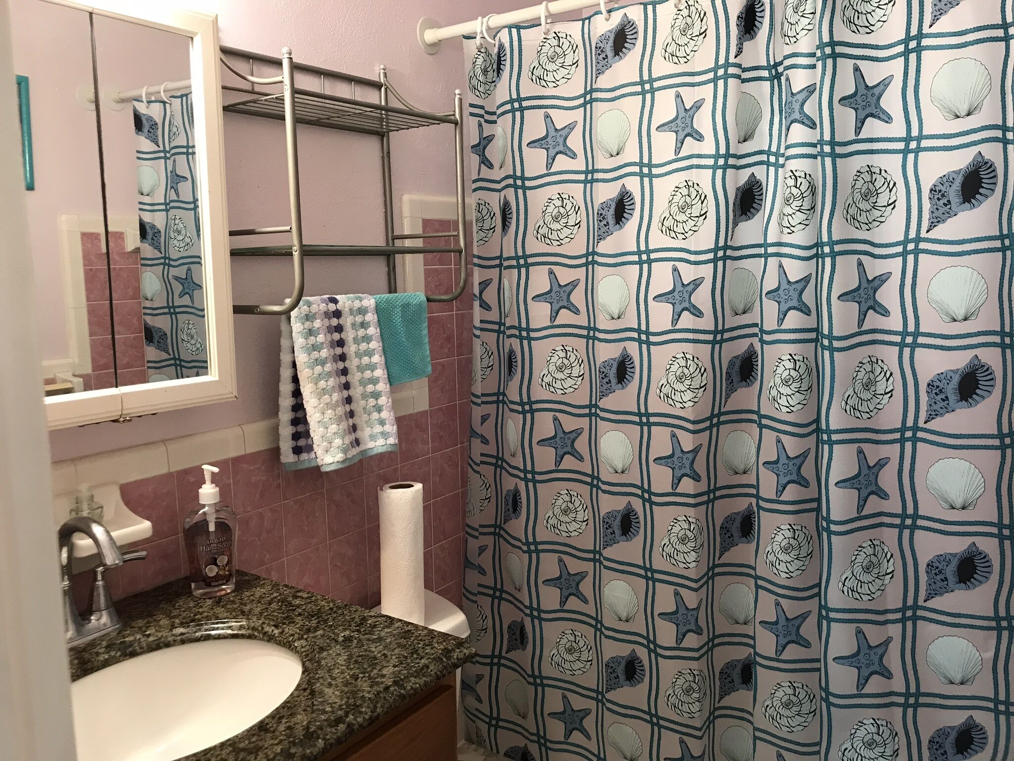 Une salle de bain équipée d’un lavabo, d’un miroir, d’un porte-serviettes et d’un rideau de douche orné de motifs d’étoiles de mer et de coquillages.