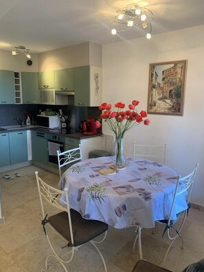 Un coin repas avec une table dressée pour quatre personnes, un vase rempli de fleurs rouges et, au fond, une cuisine.