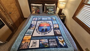 Ein Schlafzimmer mit einem Bett, das mit einer Patchworkdecke mit See- und Familienthemen bezogen ist, einem hölzernen Kopfteil, einer Nachttischlampe und einem Fenster mit Jalousien.