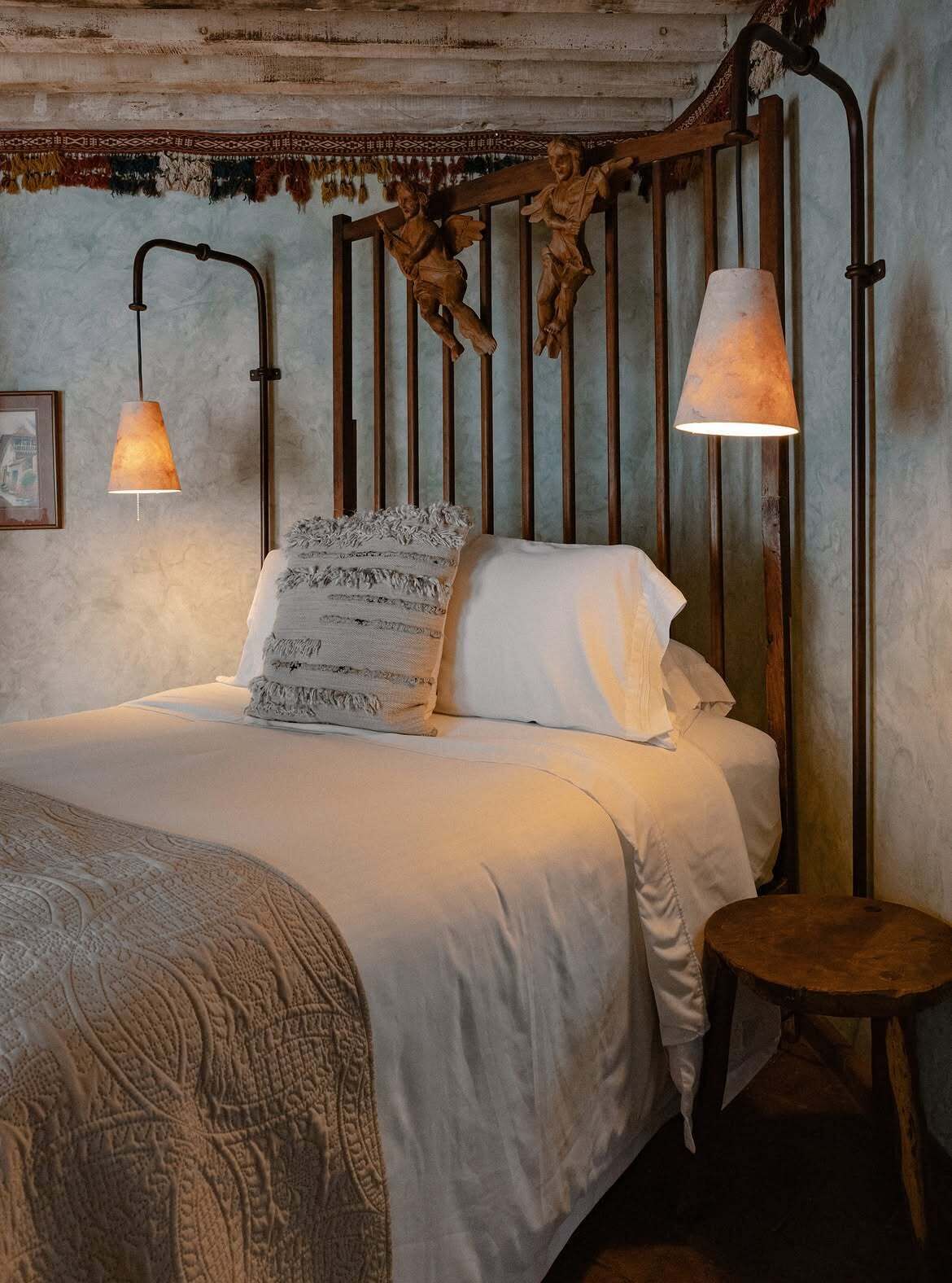 Een bed met hoofdbord, twee bedlampen, een gebreid plaid en een klein houten krukje.