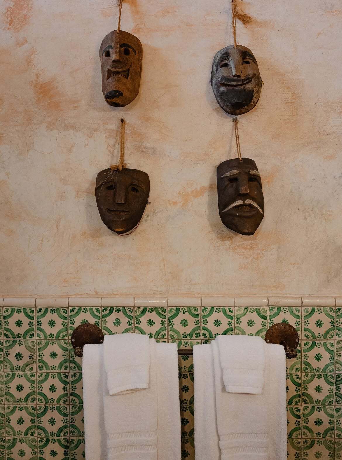A bathroom with four wooden masks hanging on the wall, a tiled floor with a green and beige pattern, and two white towels on a rack.