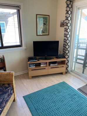 Un salon avec un téléviseur à écran plat posé sur un meuble en bois, un fauteuil en osier, un tapis turquoise et une porte-fenêtre coulissante agrémentée d’un rideau à motifs géométriques.