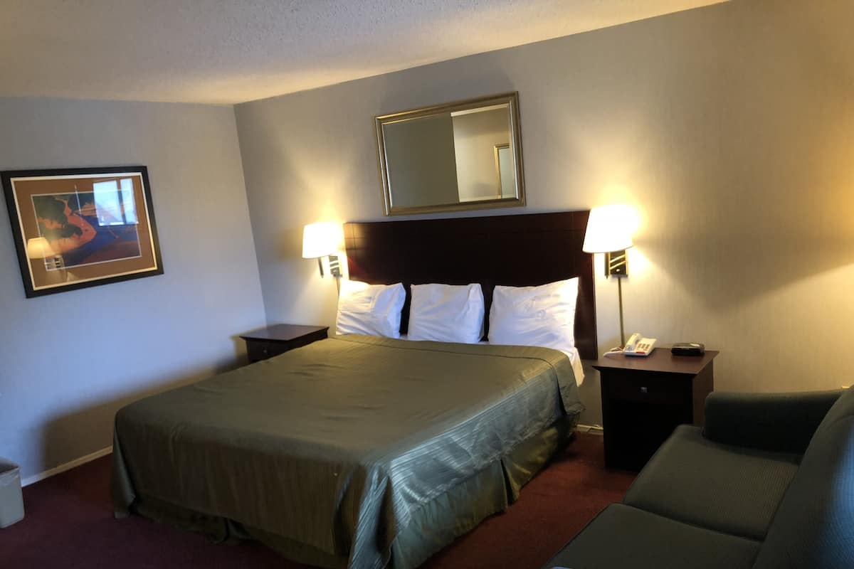 Quarto de hotel com uma cama grande, duas mesas de cabeceira com abajures, uma cadeira, um quadro na parede e um espelho sobre a cama.