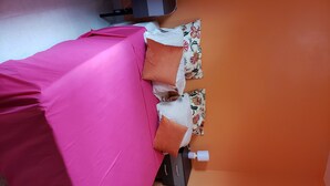 A bed with a pink bedspread and orange pillows, a white pillow with a floral pattern, and a small white object on the side.