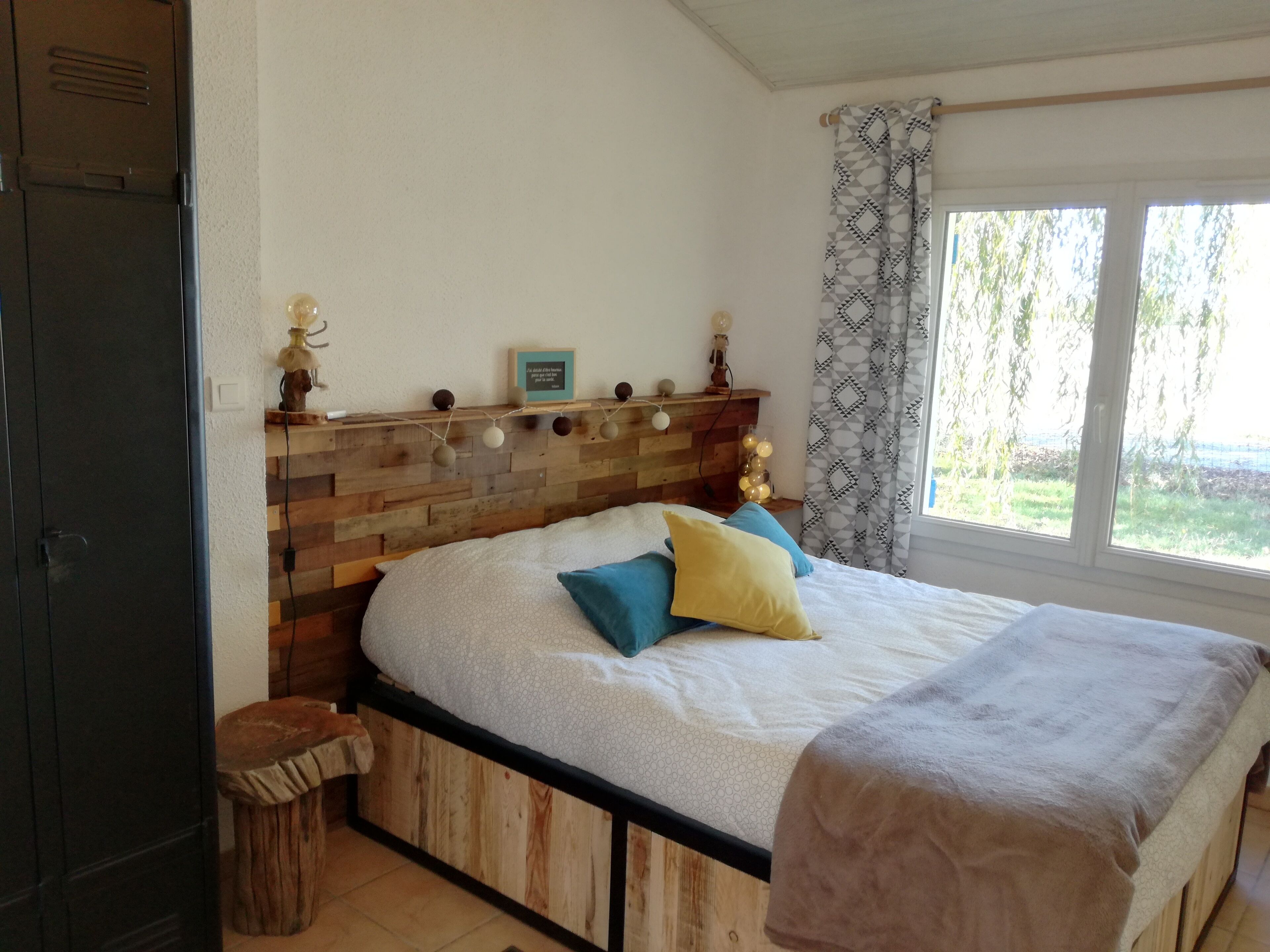 Une chambre à coucher avec une tête de lit en bois, un lit recouvert d’une couette blanche à motifs, des oreillers bleus et jaunes, et une fenêtre donnant sur un paysage verdoyant.