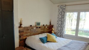 A bedroom with a wooden headboard, a bed with a white and patterned bedspread, blue and yellow pillows, and a window with a view of greenery. - A peaceful holiday 8 minutes from the beach (le perrier)