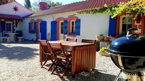 A patio area with a wooden table and chairs, a barbecue grill, and a building with red brick walls and blue shutters. - A peaceful holiday 8 minutes from the beach (le perrier)