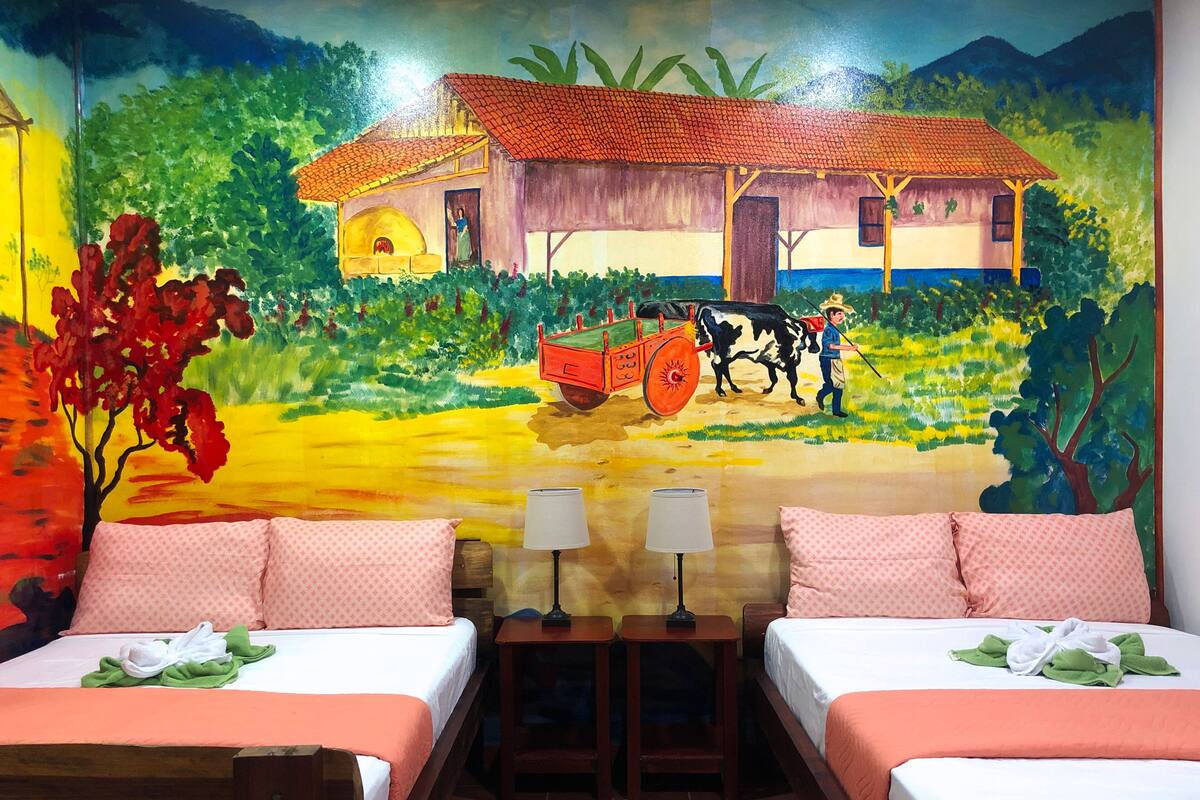 A hotel room with two beds, a mural of a rural scene, and a small table with a lamp between the beds.
