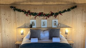A cozy bedroom with a wooden headboard, a bed with white linens, two pillows, and a dark blue throw blanket. There are two bedside tables with lamps, and a garland with red berries and pinecones above the headboard. - Luxury and Peaceful 1-bed Roundhouse With Hot Tub (Glasgow)