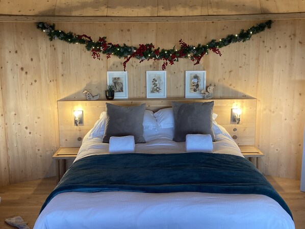 Een gezellig slaapkamer met een houten hoofdbord, een bed met witte lakens, twee kussens en een donkerblauwe plaid. Er staan twee nachtkastjes met lampen, en een guirlande met rode bessen en dennenappels boven het hoofdbord.