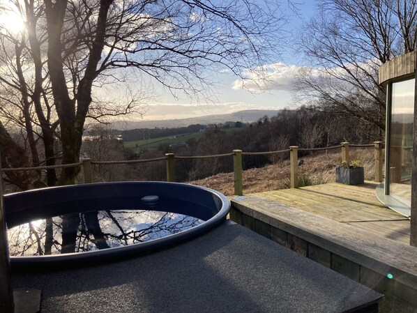 Una terrazza in legno con vasca idromassaggio, affacciata su un paesaggio panoramico con alberi e colline in lontananza.