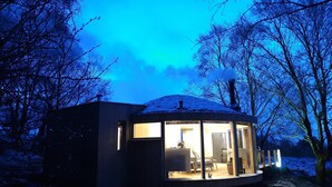 A circular, glass-enclosed cabin with a visible interior containing a sofa and a table, surrounded by snow-covered ground and trees. - Luxury and Peaceful 1-bed Roundhouse With Hot Tub (Glasgow)