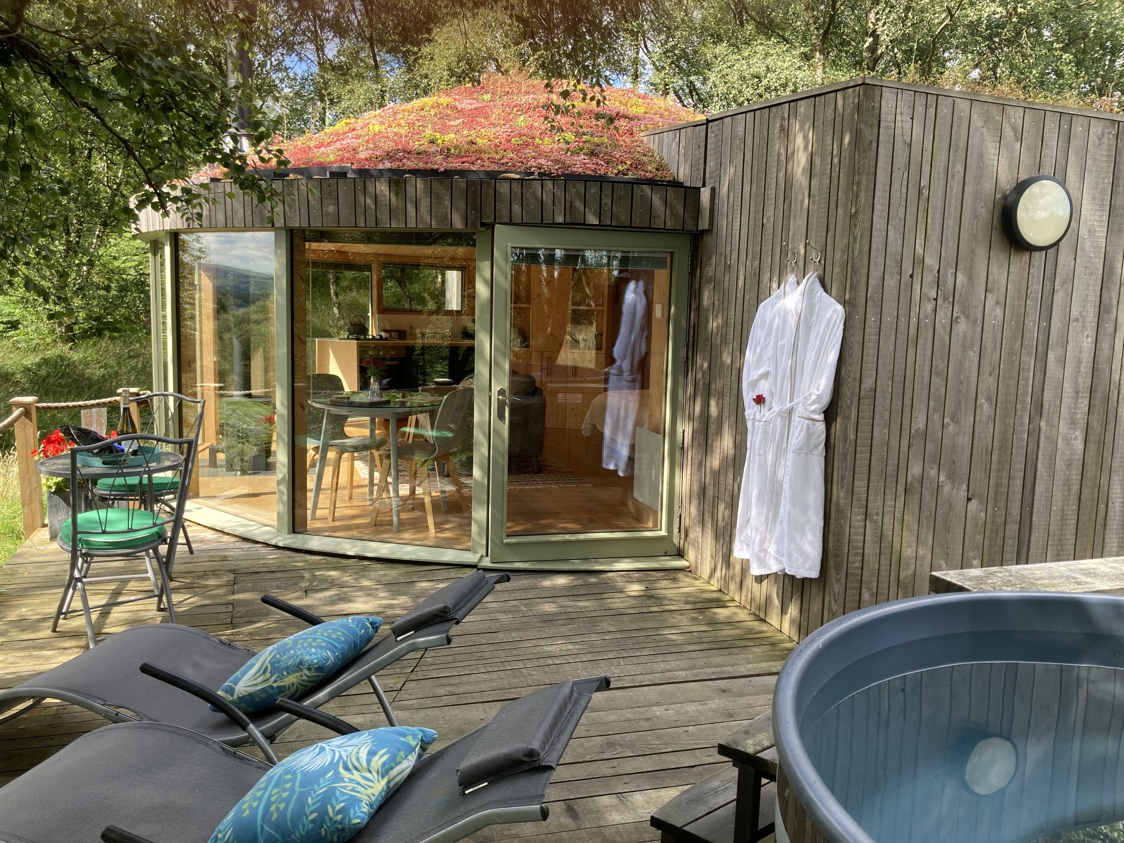 Una terraza de madera con sillones de descanso y un jacuzzi, una sala acristalada con puerta corredera, y una pared de madera con un perchero para toallas.