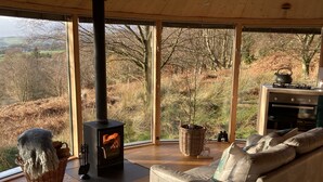 A cozy living room with a large window, a fireplace, a sofa, and a kitchen area. - Luxury and Peaceful 1-bed Roundhouse With Hot Tub (Glasgow)