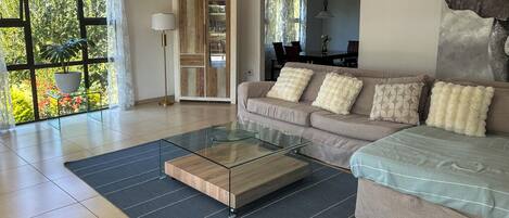 Una sala de estar moderna con una mesa de centro de vidrio, un sofá con almohadones decorativos, una estantería y vistas a zonas verdes a través de amplios ventanales.