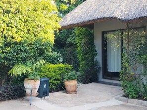 A thatched-roof building with large windows, surrounded by lush greenery and potted plants.