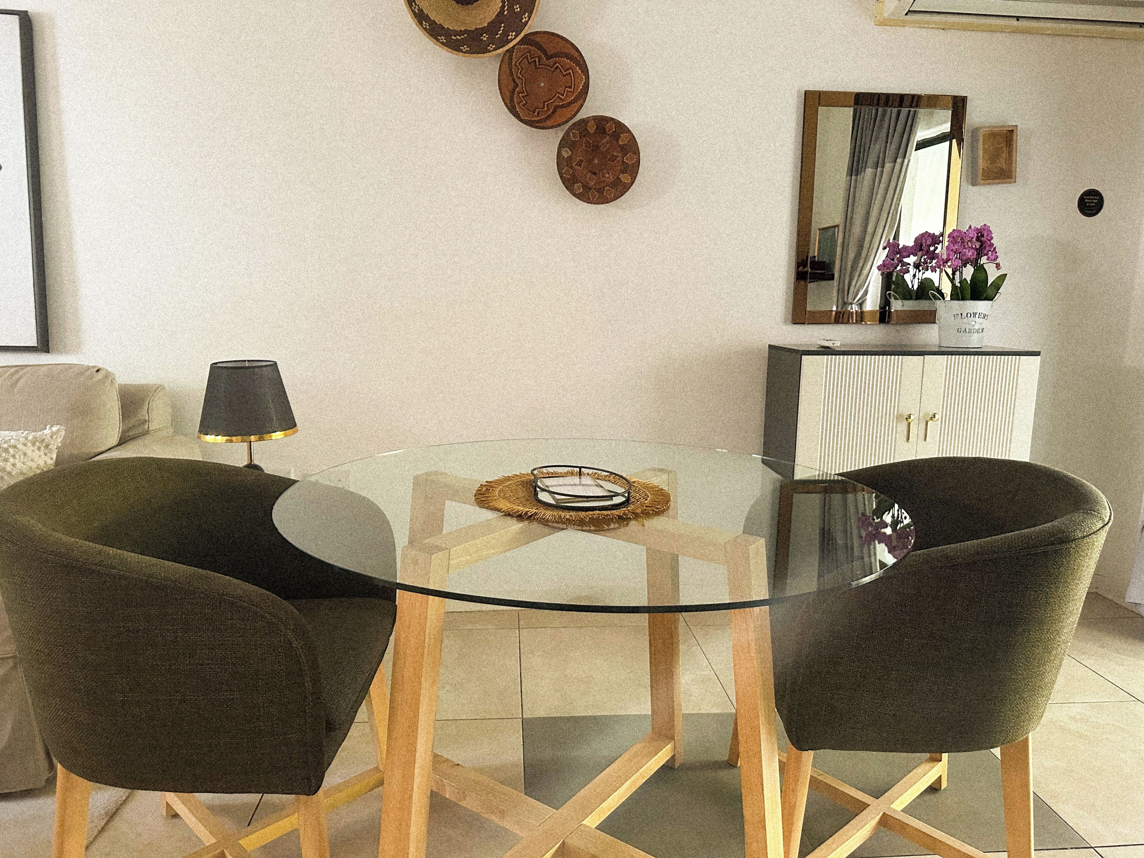 Uma sala de estar moderna com uma mesa de vidro, duas cadeiras verde-escuras, um abajur, pratos decorativos na parede, um espelho e um aparador com um vaso de flores.
