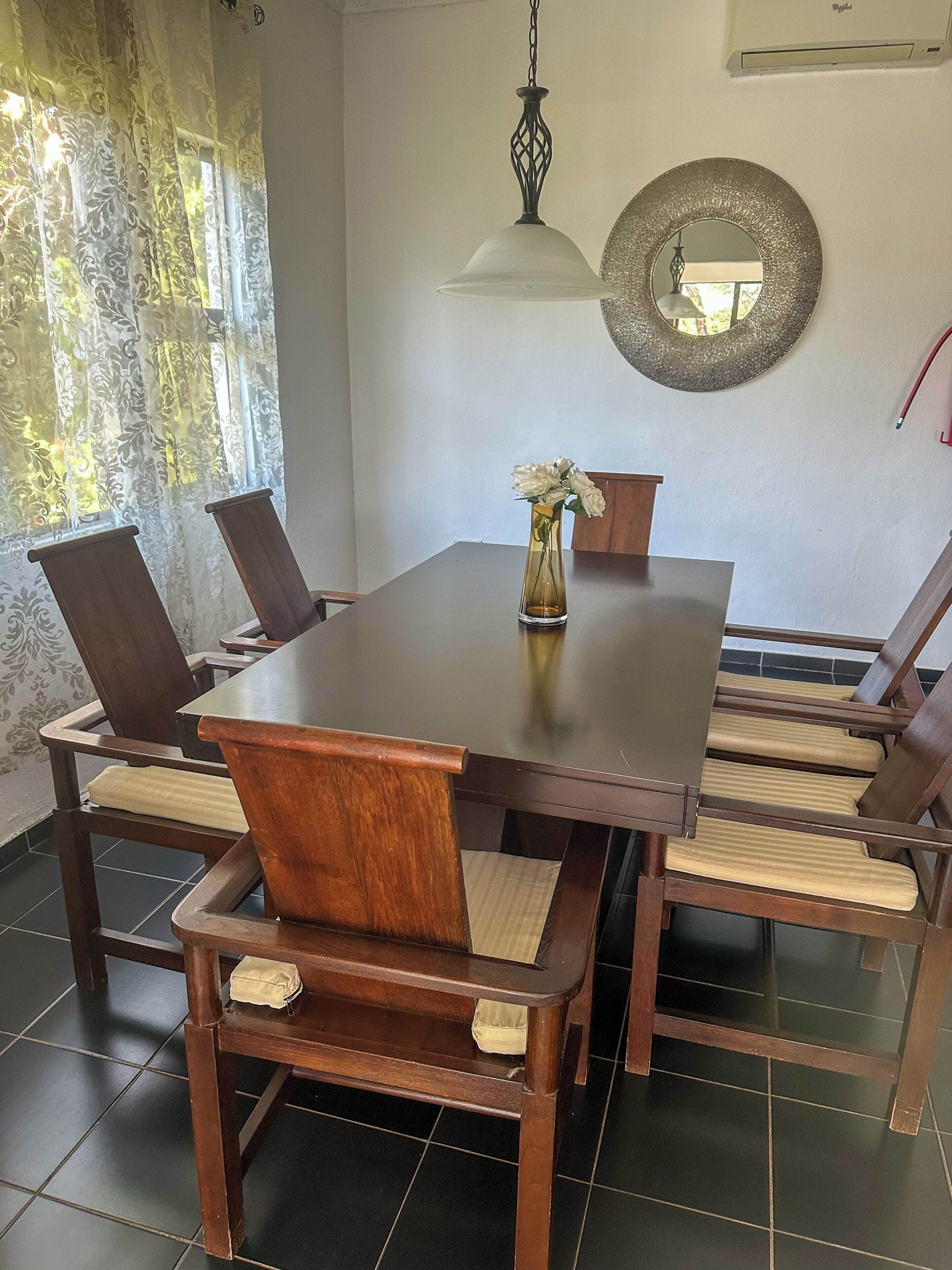 A dining area with a wooden table and chairs, a vase with flowers, a hanging light fixture, and a decorative mirror on the wall.