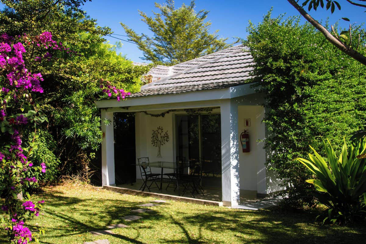 Un pequeño edificio de una planta, de color blanco, con techo de tejas, puerta de vidrio y un patio con mobiliario exterior.