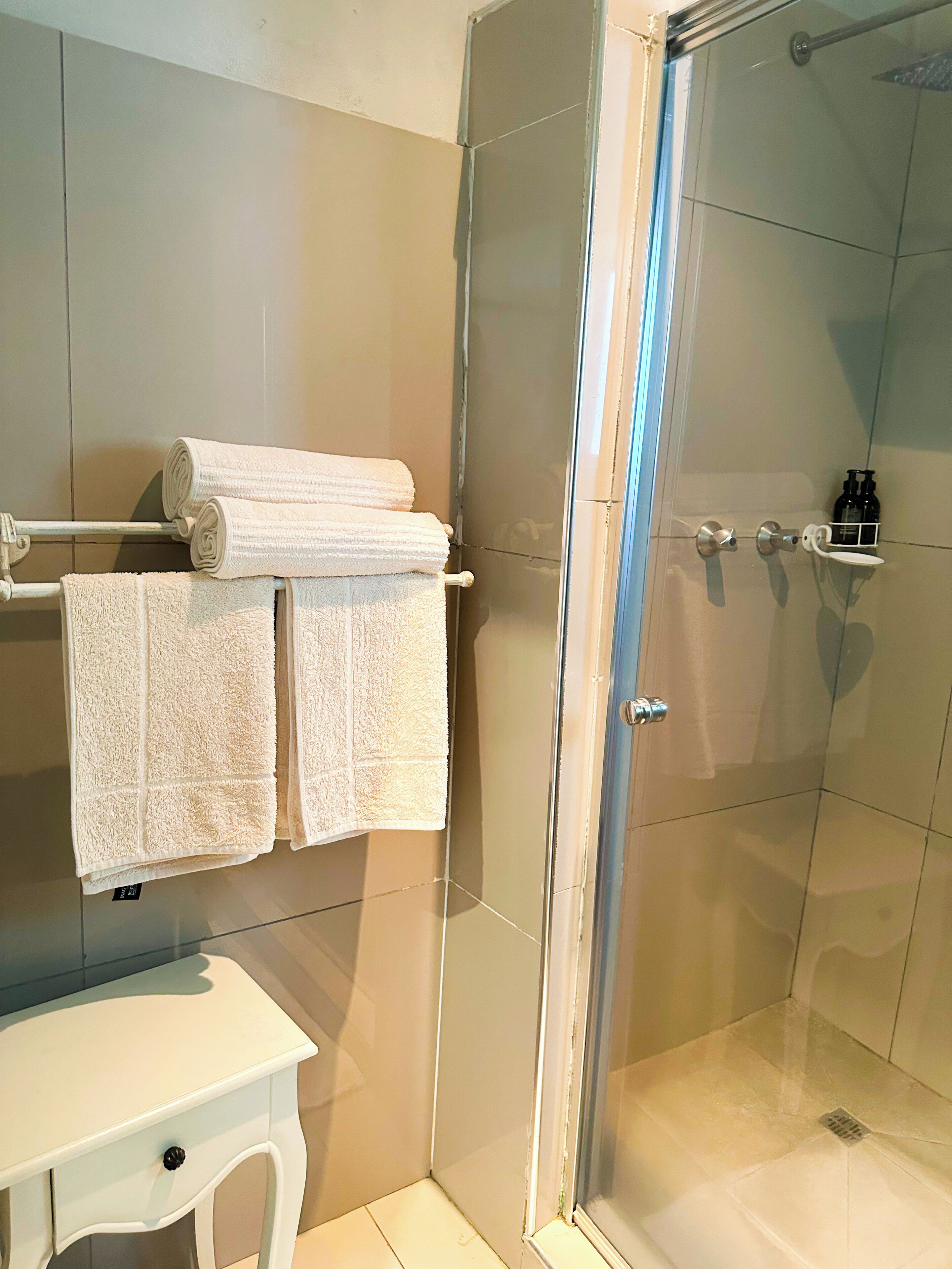 Un baño con cabina de ducha de vidrio, un perchero con toallas dobladas, un lavabo y un estante pequeño con artículos de aseo.