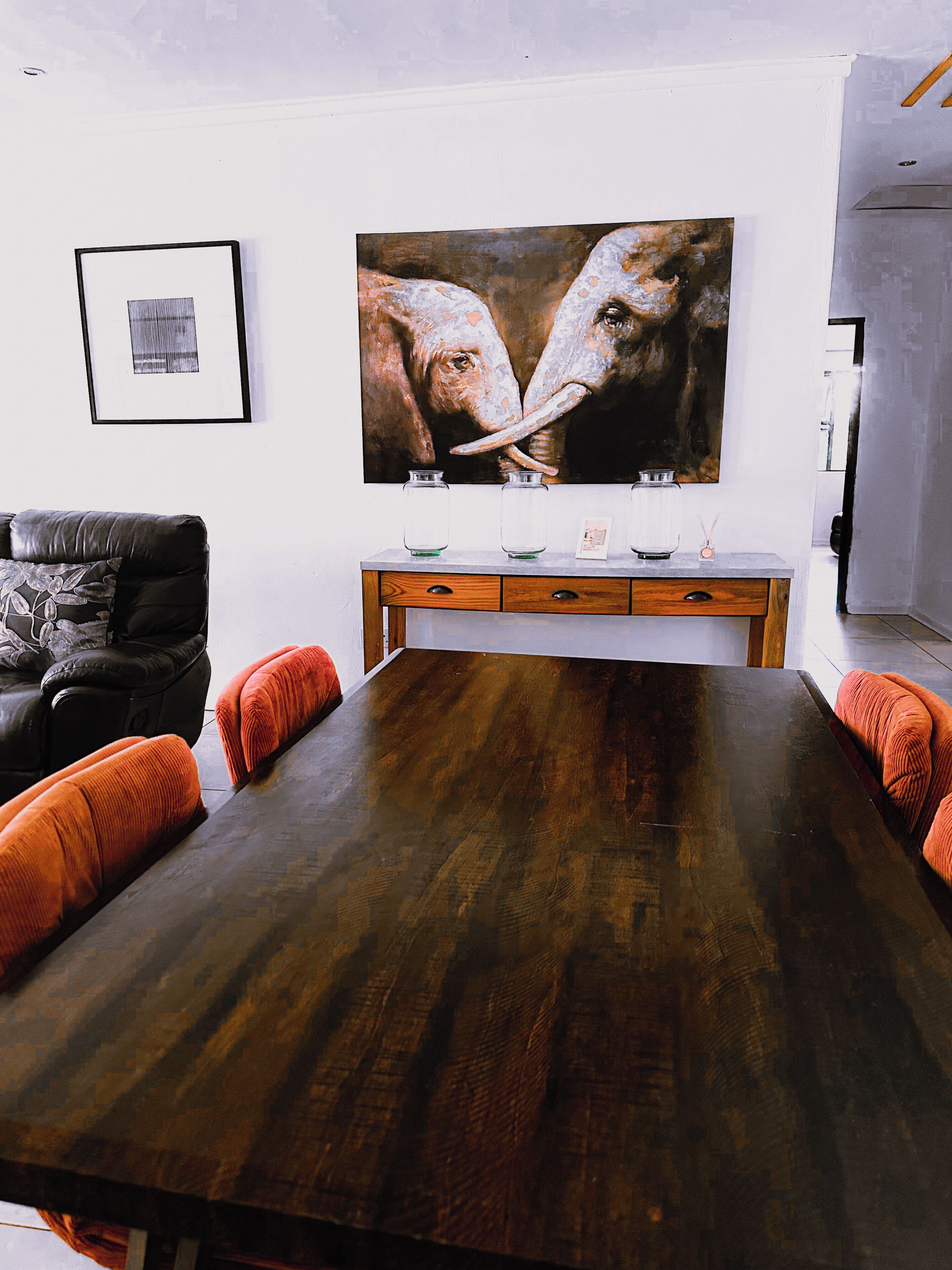 Un coin repas avec une table en bois, des coussins orange, un tableau encadré et une grande peinture au mur.