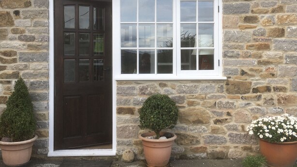 A stone building with a dark wooden door and a white-framed window. There are two potted plants, one with a small tree and another with white flowers, placed on the ground. - Groom's Cottage  a fabulously quiet paradise in the heart of the Peak District (Castleton)