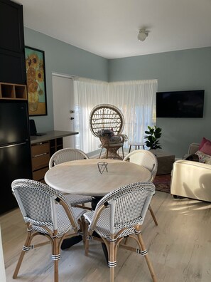 Un coin repas avec une table ronde et des chaises en osier, un téléviseur fixé au mur et une grande fenêtre avec des rideaux.