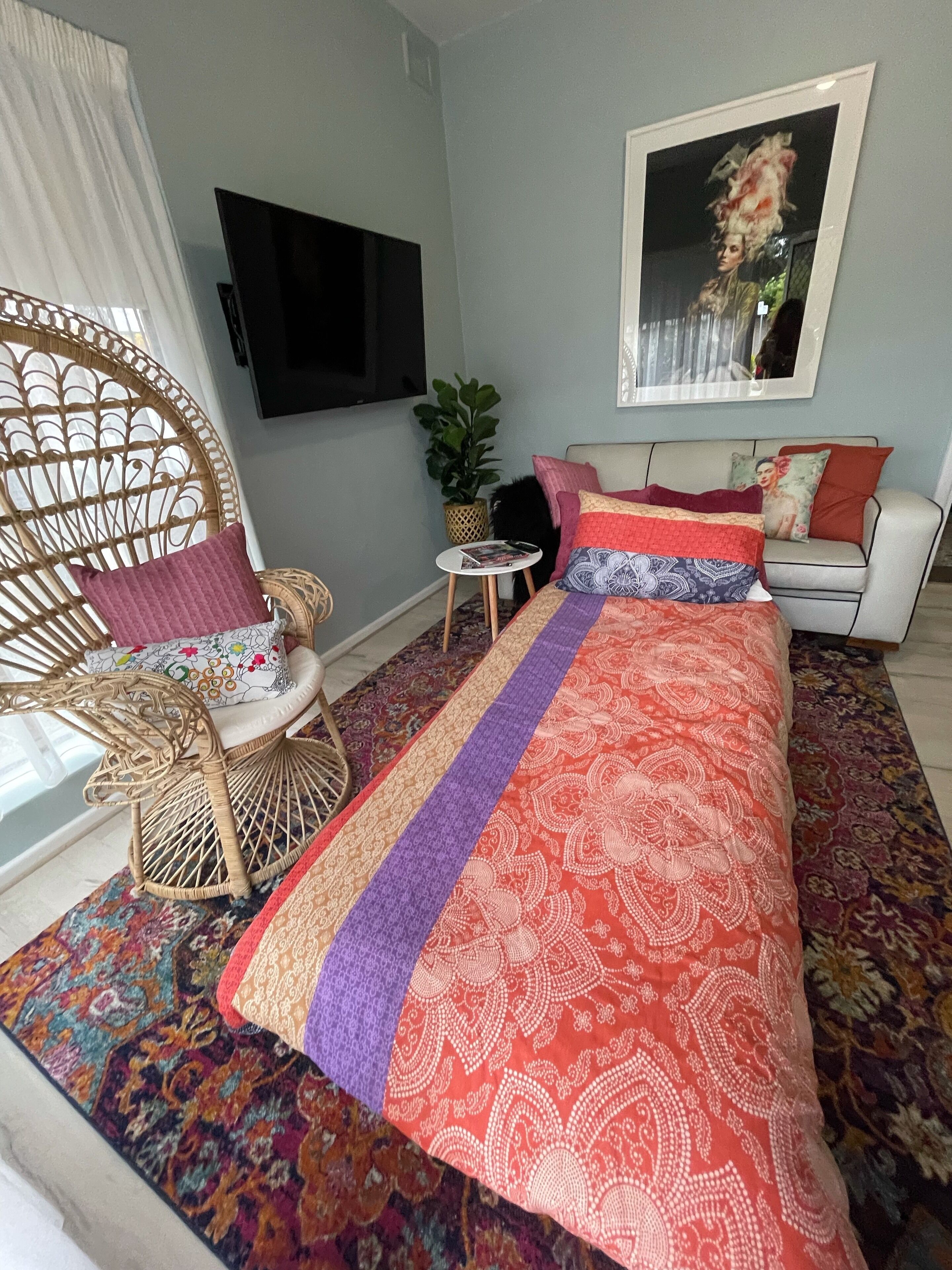 Een slaapkamer met een groot bed, een rieten stoel, een klein tafeltje en een televisie aan de muur.