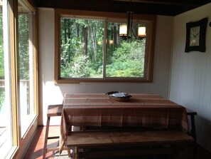 A room with a wooden table and bench, a window with a view of a forest, and a wall-mounted clock.