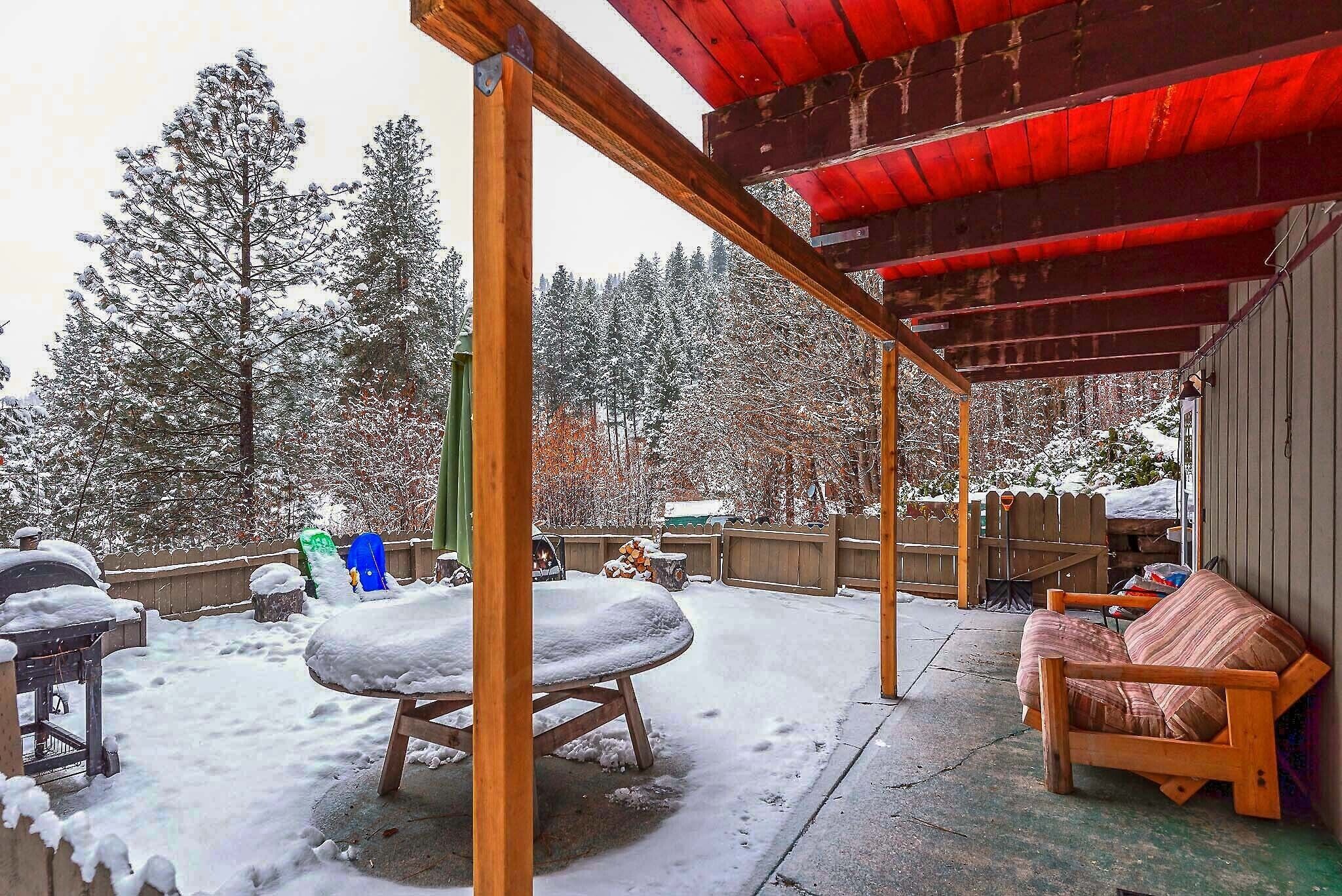 A covered outdoor area with a wooden bench and a table covered in snow, surrounded by snow-covered trees and a wooden fence.