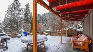 A covered outdoor area with a wooden bench and a table covered in snow, surrounded by snow-covered trees and a wooden fence.