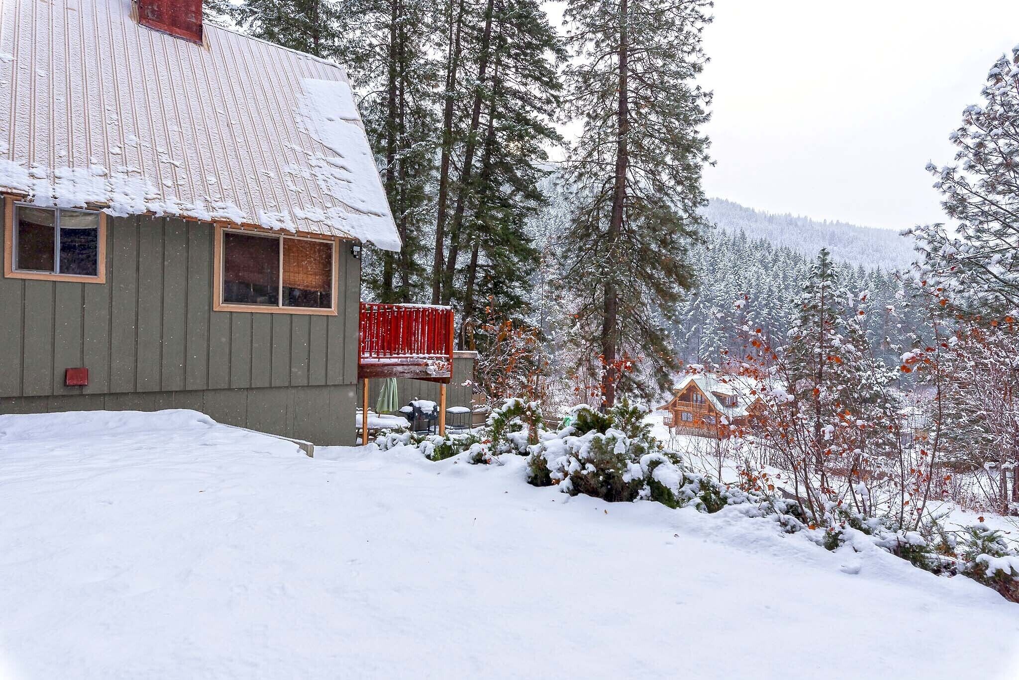 A house with a metal roof and a red balcony, surrounded by snow-covered trees and a forested hillside.