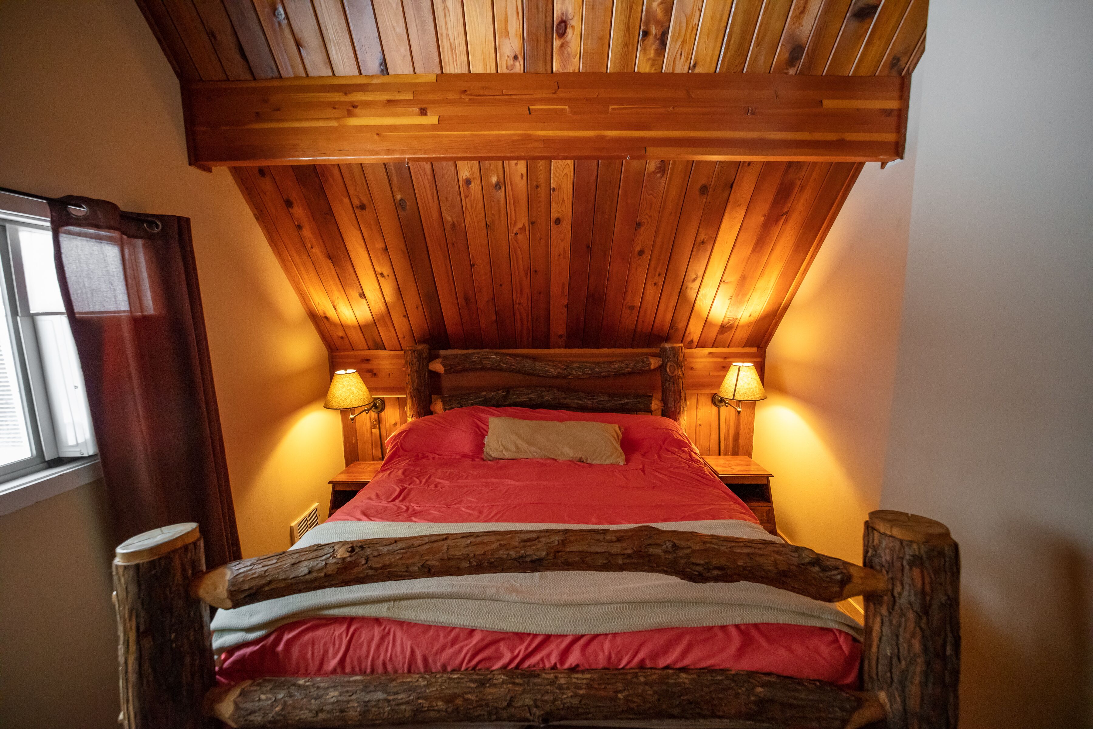 Ein Bett aus Holz mit einer rot-weißen Matratze, zwei Nachttischlampen und einem Fenster mit Vorhängen.
