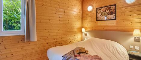A wooden-paneled room with a bed, a window with curtains, a wall-mounted lamp, and a framed picture.