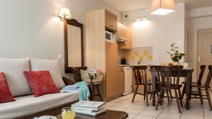 A living room with a sofa, a coffee table with a book and glasses, and a dining area with a table and chairs. - Appart Cosy le Long de la Route des Vins d'Alsace (Bergheim)